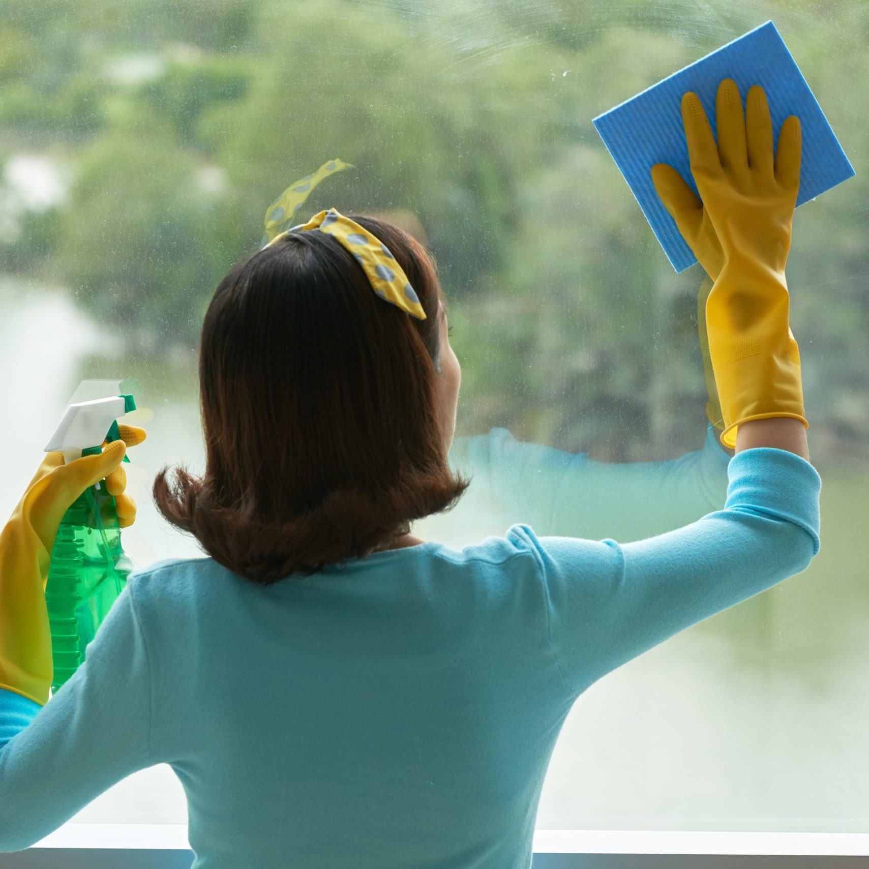 Back view of unrecognizable housekeeper cleaning panoramic window with help of rag and glass cleaner, picturesque view on background